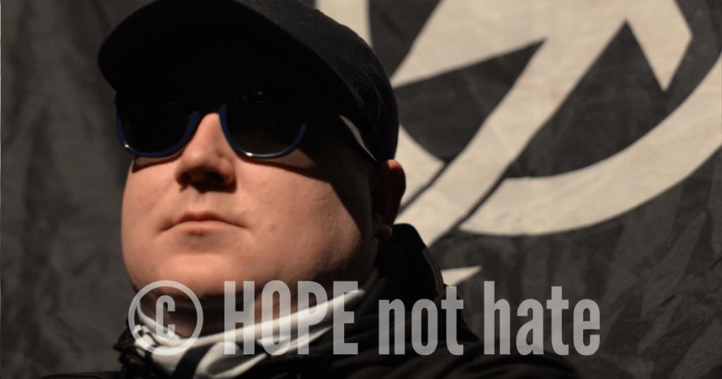 a picture of HOPE not hate informant Robbie Mullen, wearing National Action gear with the National Action symbol in the background