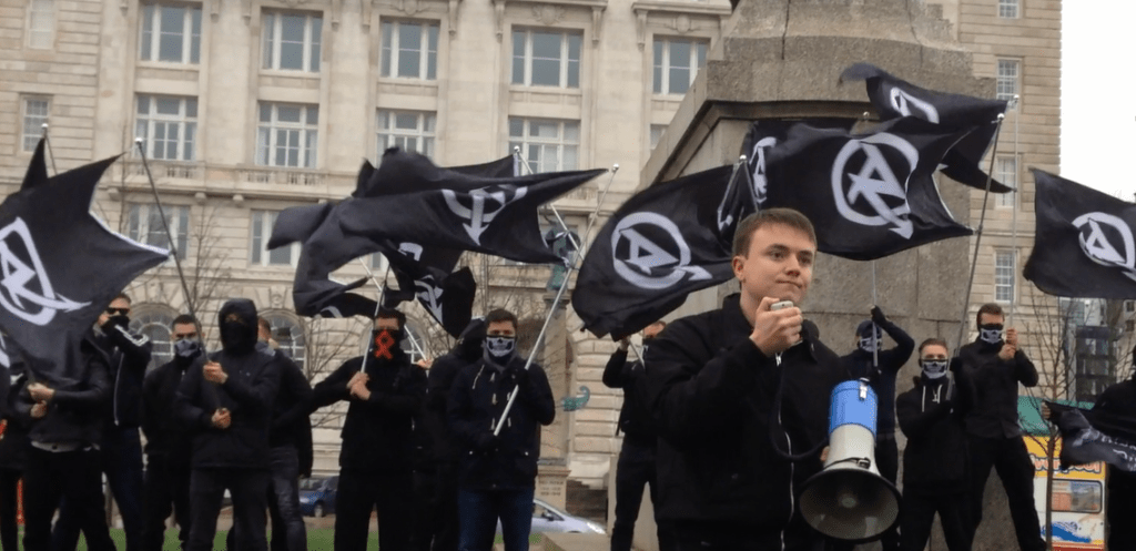 National Action member Jack Renshaw is holding a megaphone at a National Action demonstration. There are many members of National Action behind him, all dressed in black, covering their faces and waving the national action flag