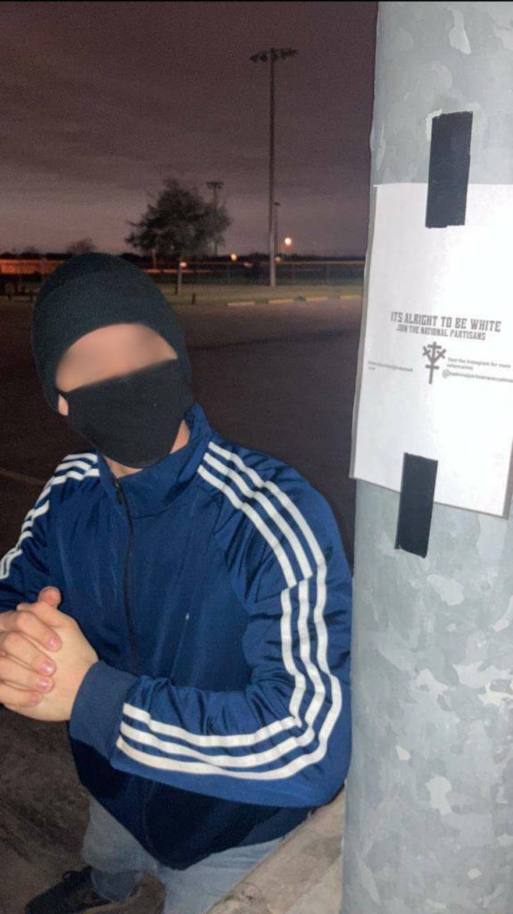 A teenager with his face blurred out is posing next to graffiti that says "It's alright to be white. Join the National Partisans"
