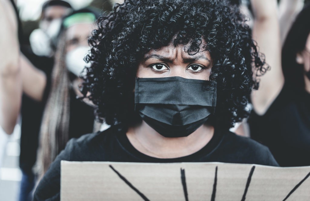 A woman is wearing a mask, staring directly into the camera. She is holding a placard. 